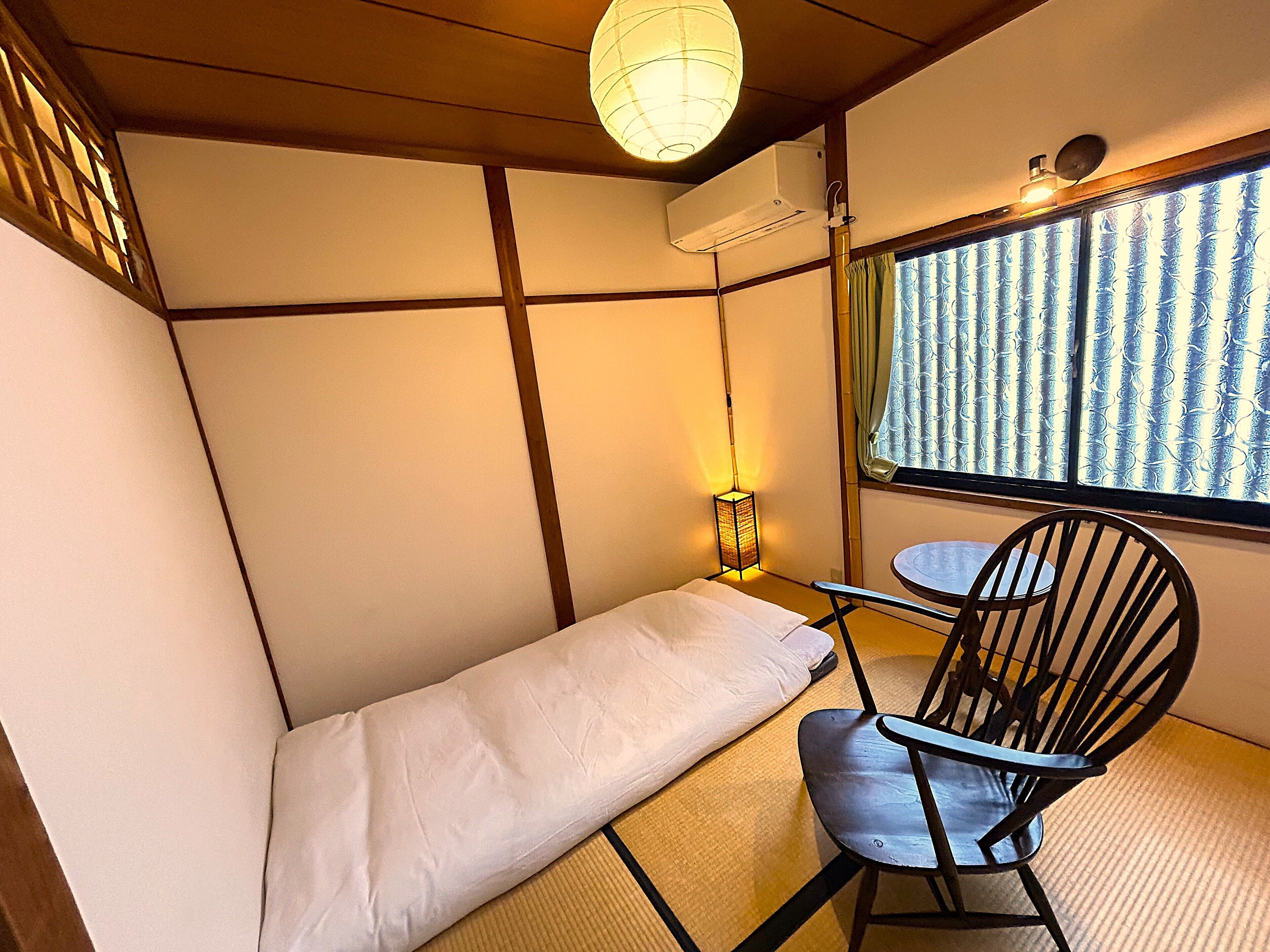 Ein Zimmer mit Tatami-Boden, einem niedrigen Bett, einem Stuhl, einem kleinen Tisch, einem Fenster mit gemusterten Vorhängen und einer hängenden Papierlaterne.