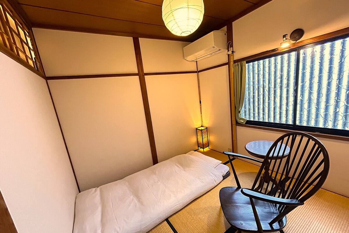 Una habitación con suelo de tatami, una mesa baja, una silla, una cama con colchón blanco, un farolillo de papel colgante y una ventana con cortinas estampadas.