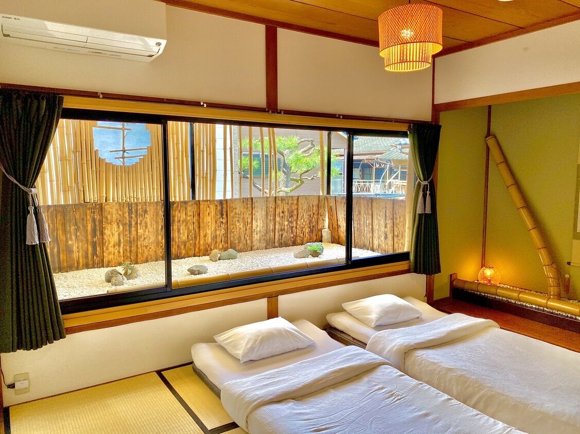 Una habitación con dos camas, un ventanal con vistas a una valla de bambú y rocas, y una lámpara tradicional japonesa.