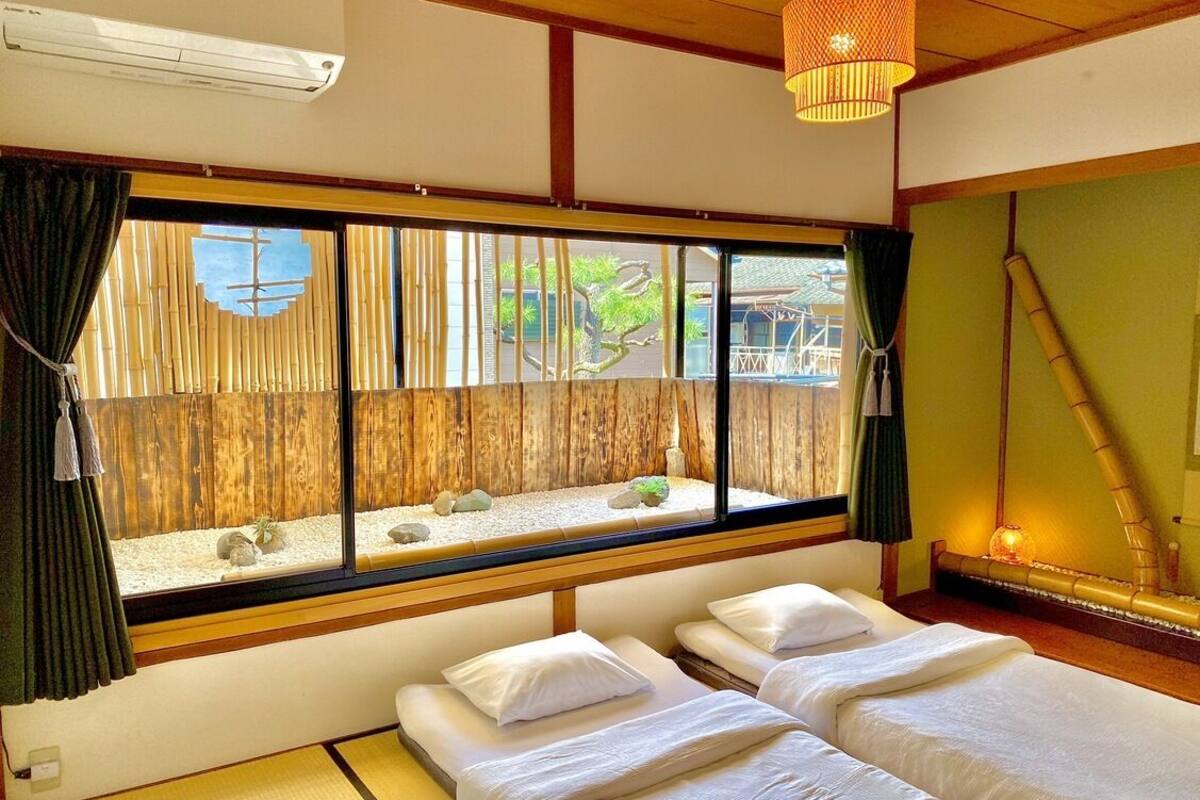 Una habitación con dos camas, un ventanal con vistas a una valla de bambú y rocas, y una lámpara tradicional japonesa.