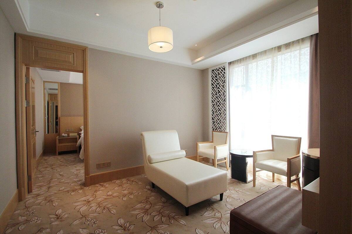 A hotel room with a bed, a sitting area with chairs, a small table, a decorative wall panel, and a large window with curtains.