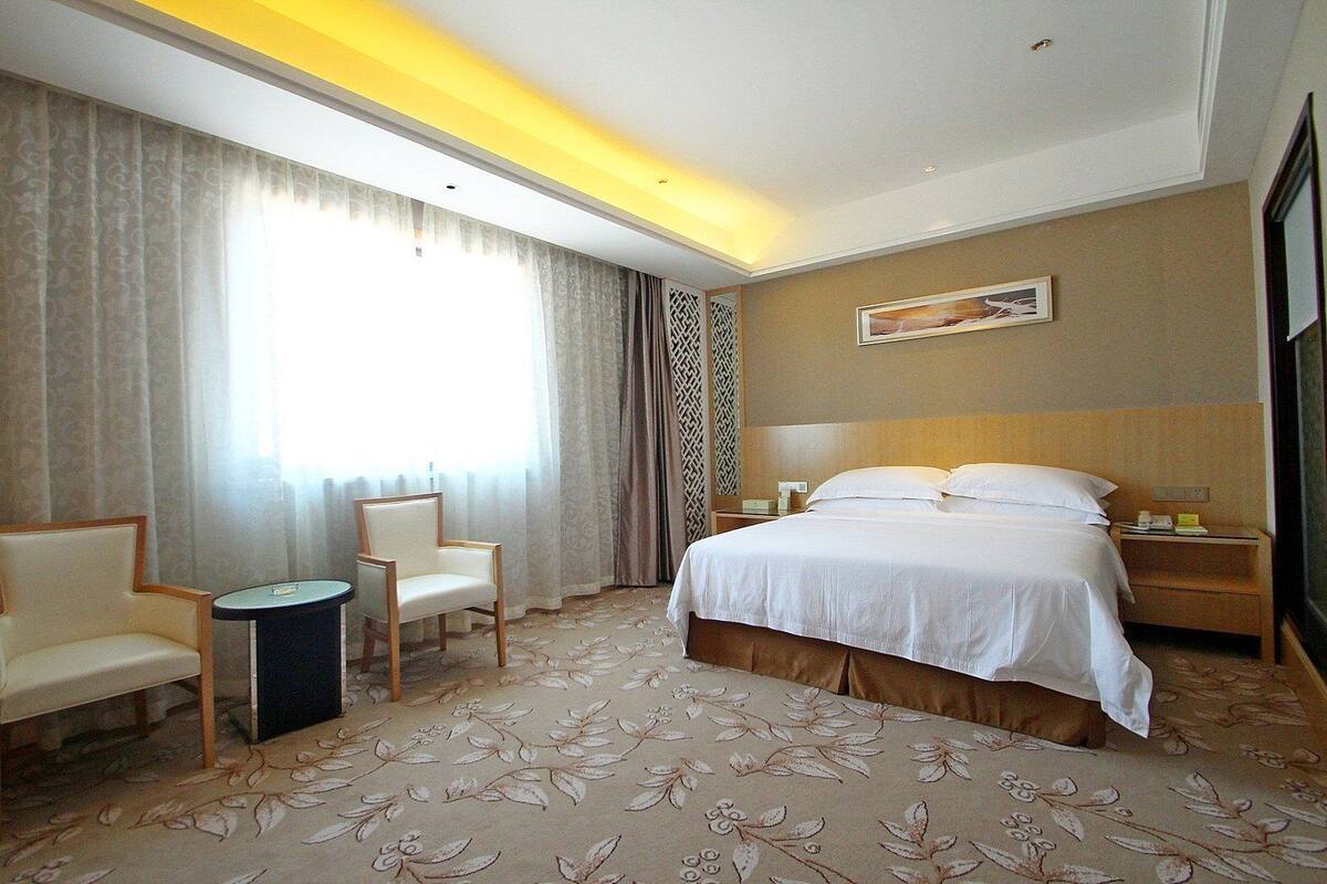 A hotel room with a bed, two chairs, a small table, a nightstand, and a large window with curtains.