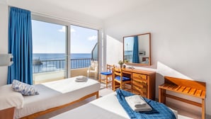 A hotel room with two beds, a desk, a chair, and a large window with a view of the sea. - Hostal Port Corona (Capdepera)