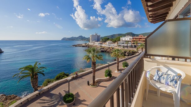 A balcony with a view of the sea, a paved walkway with palm trees, and buildings in the background. - Hostal Port Corona (Capdepera)
