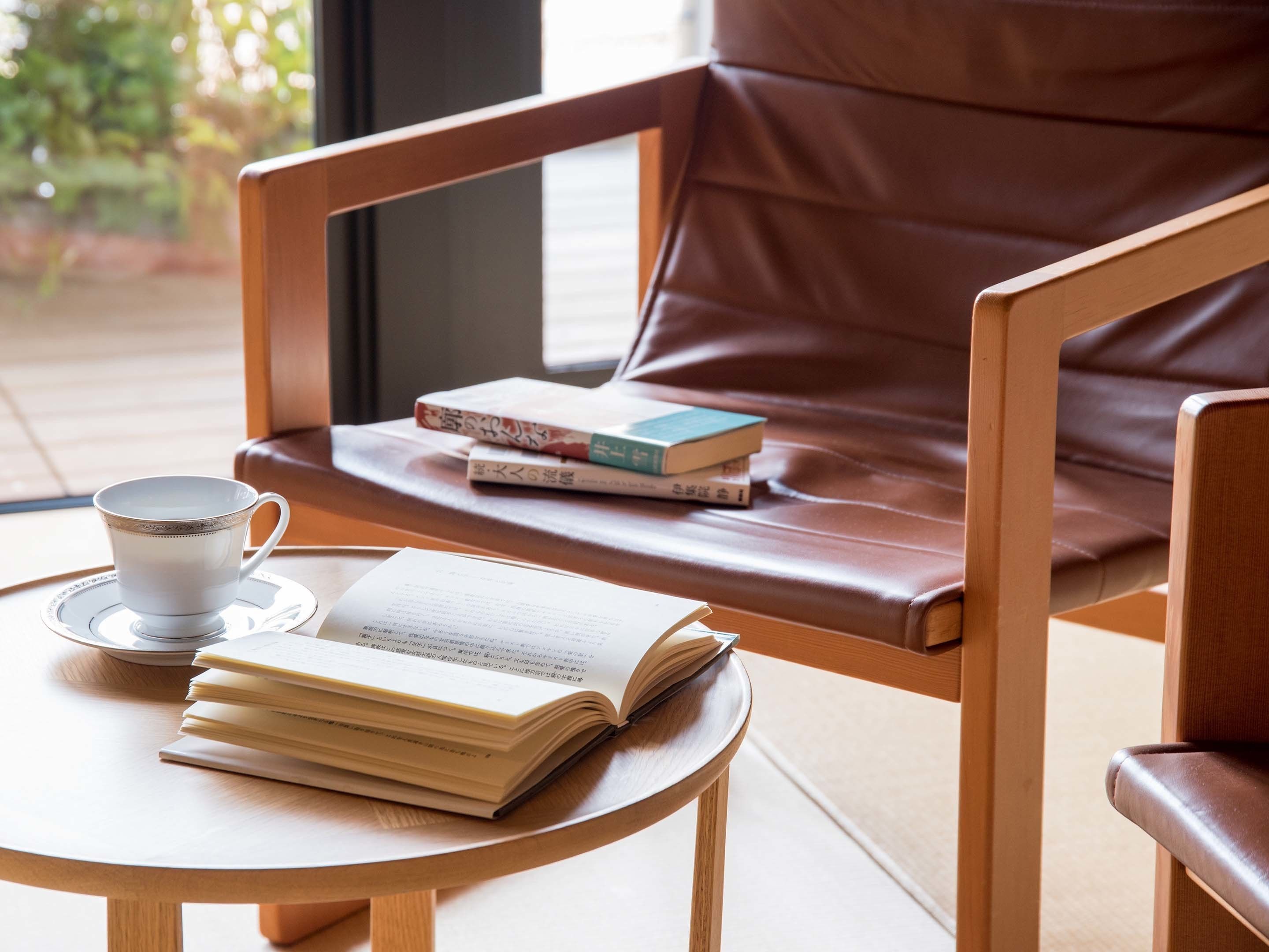 A wooden chair with a leather cushion, a small round table with a white cup and saucer, and a stack of books.