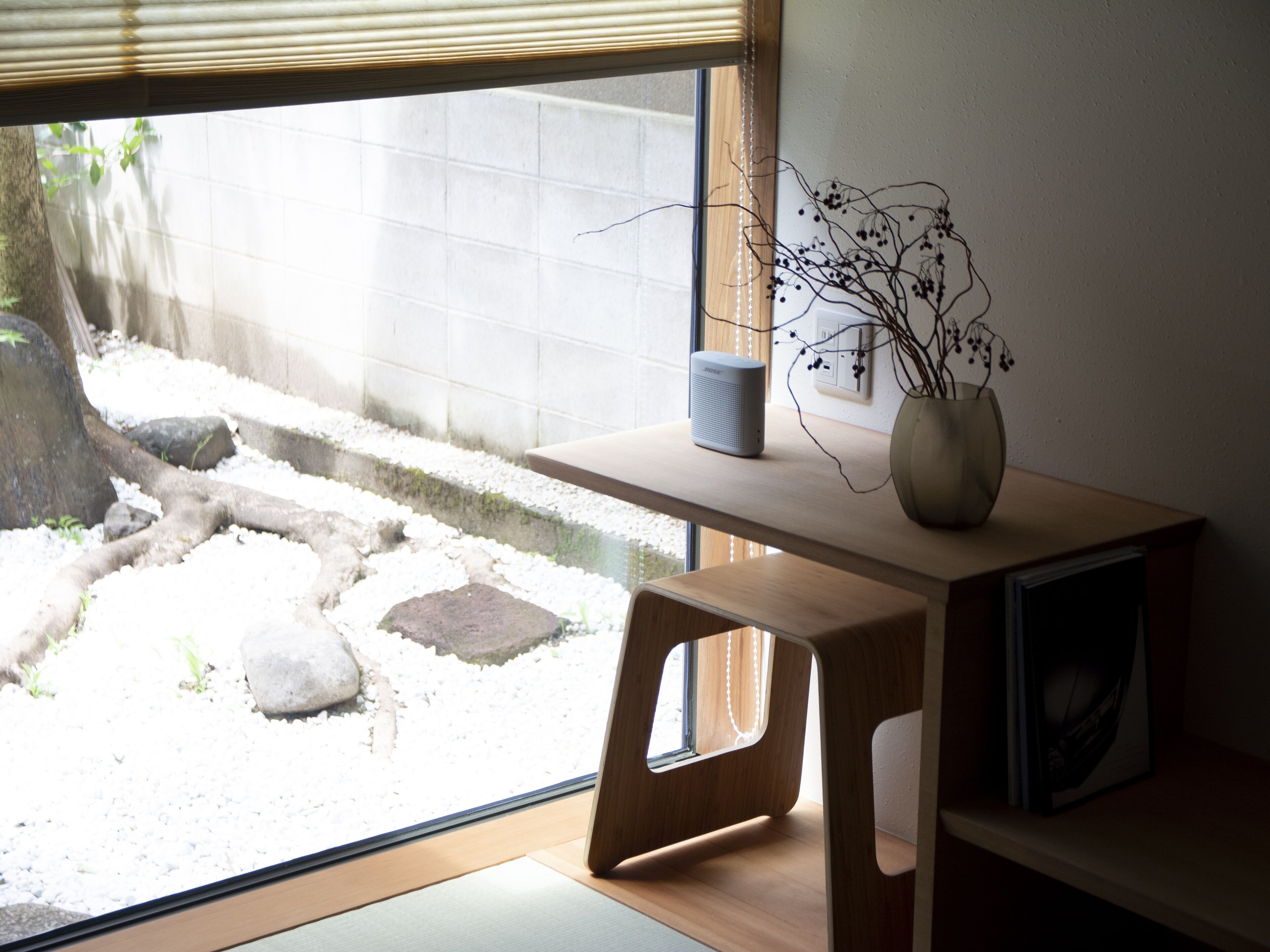 A room with a wooden desk, a vase with branches, and a window overlooking a garden.
