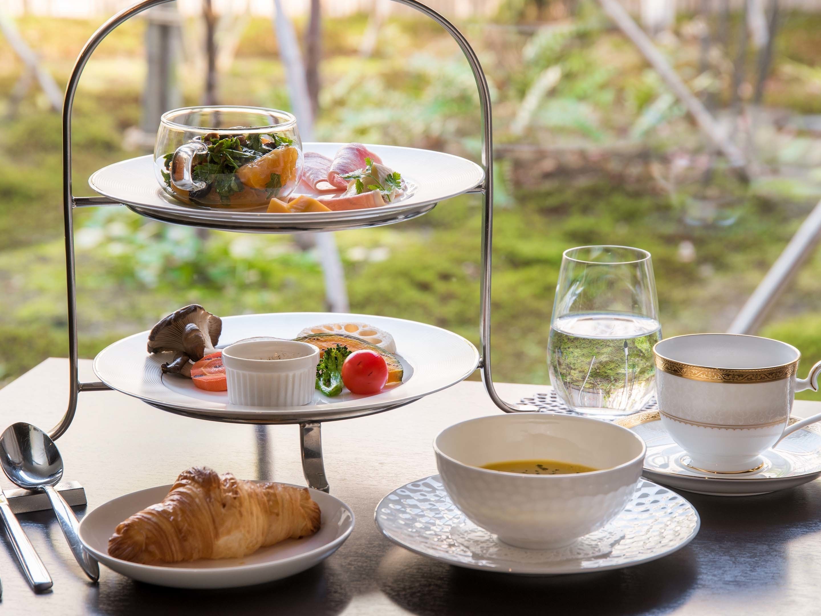 Une table extérieure à trois niveaux, dressée avec des assiettes de nourriture, un verre d’eau et une tasse, le tout mis en valeur par un décor naturel.