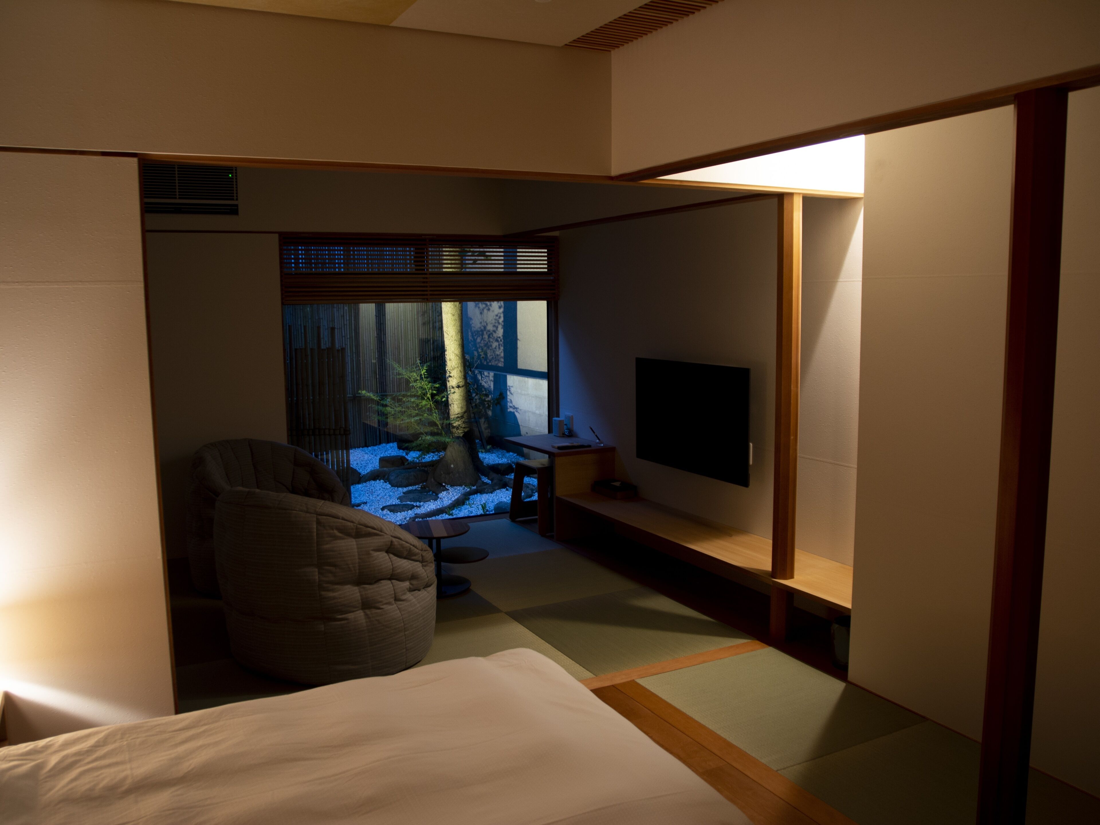 A room with a bed, a seating area with a chair, a television, and a view of a garden through a window.