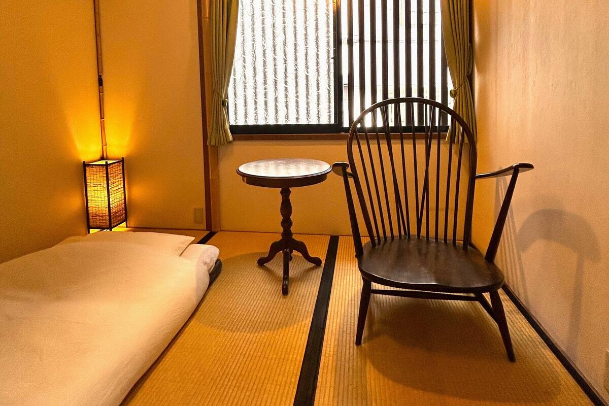 Une pièce comprenant un lit, une chaise, une petite table et une fenêtre avec des rideaux.