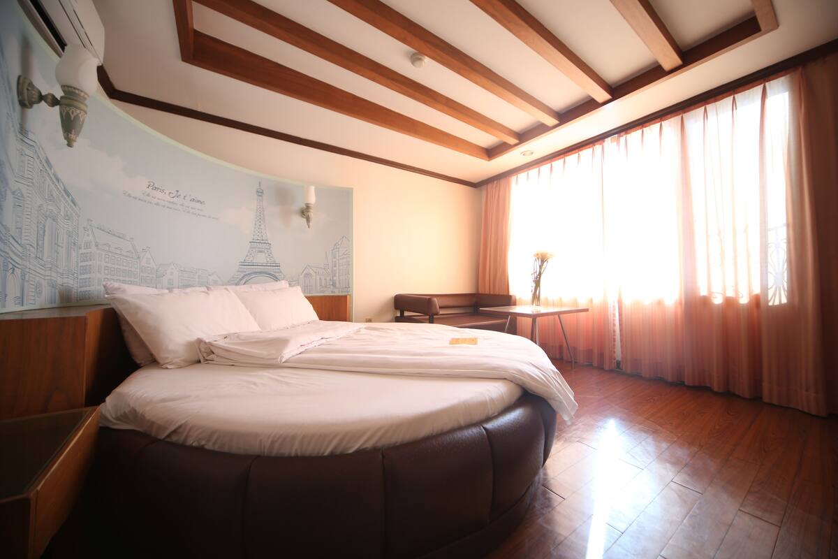 A bedroom with a large bed, a wooden ceiling, a wall mural of Paris, and a large window with curtains.