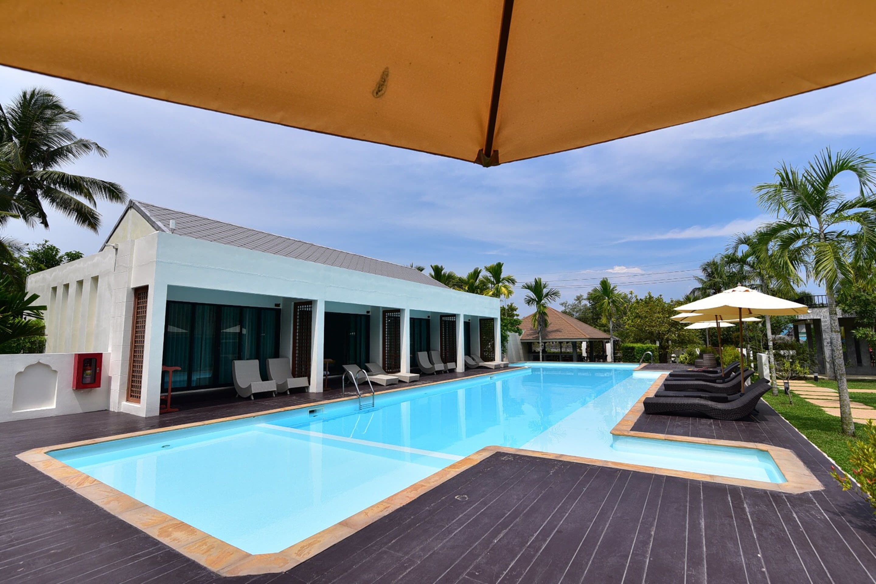 A swimming pool with lounge chairs and umbrellas, surrounded by palm trees and a building with large windows.
