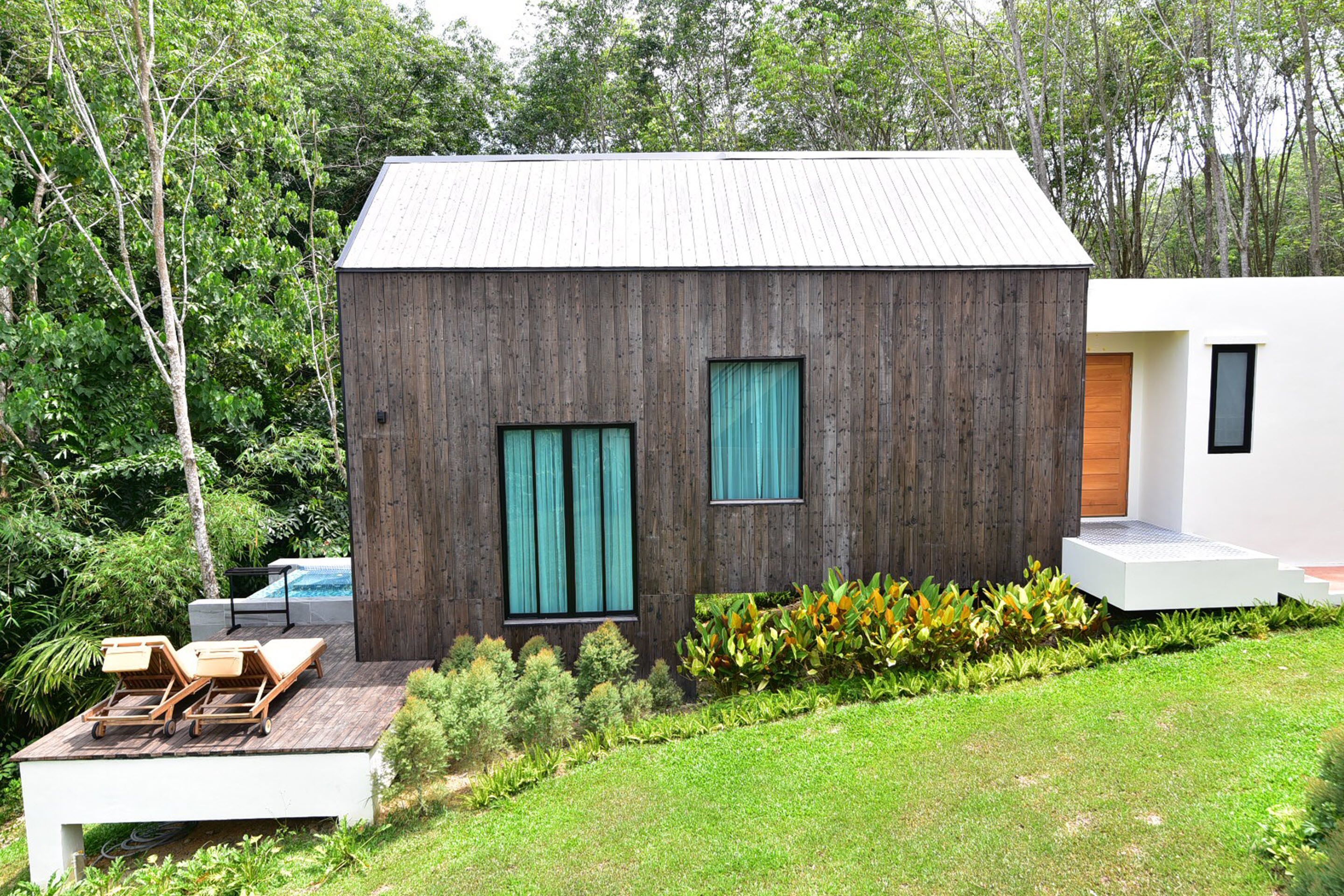A modern house with a wooden exterior, a small deck with lounge chairs, and a swimming pool in the background.