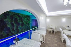 Um restaurante com mesas e cadeiras brancas, uma parede de aquário grande e piso de madeira.