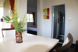 A room with a white table, a brown vase with a plant, a bed with white bedding, and a door leading to another room.