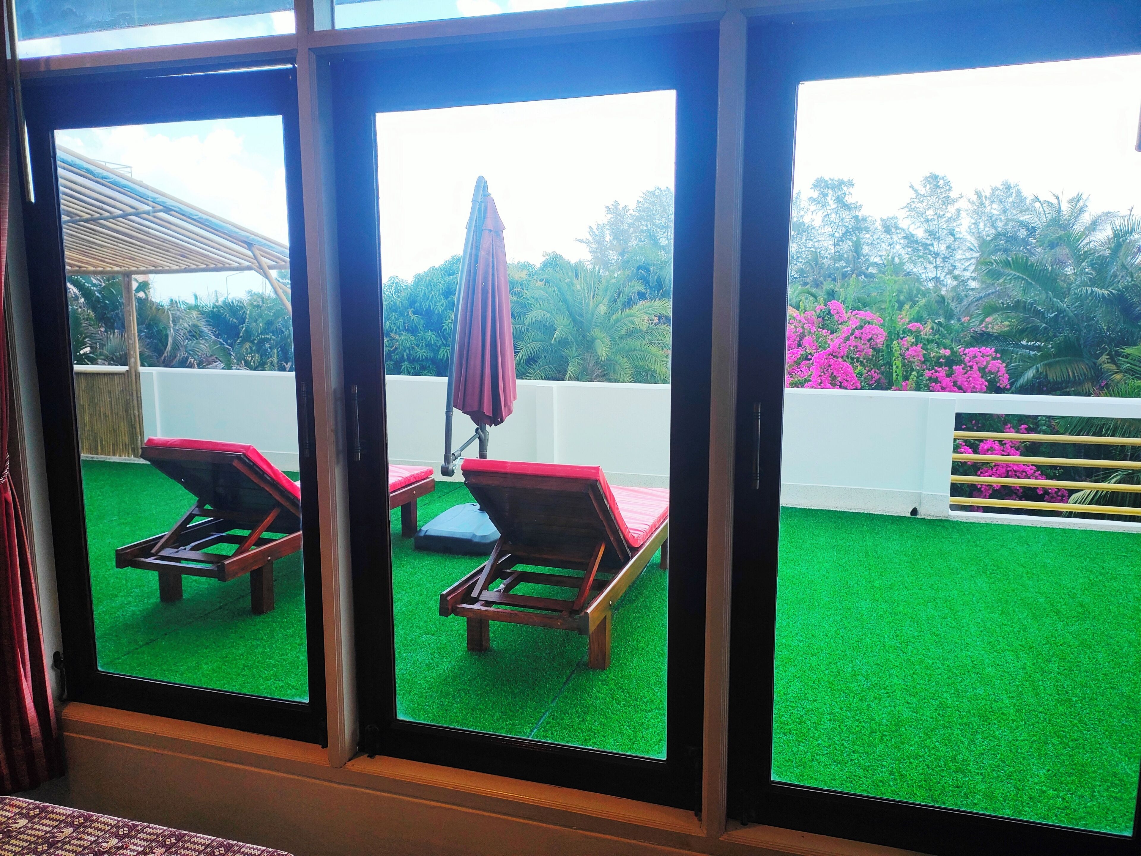 A room with a view of a green artificial lawn, two wooden lounge chairs, and a red umbrella.