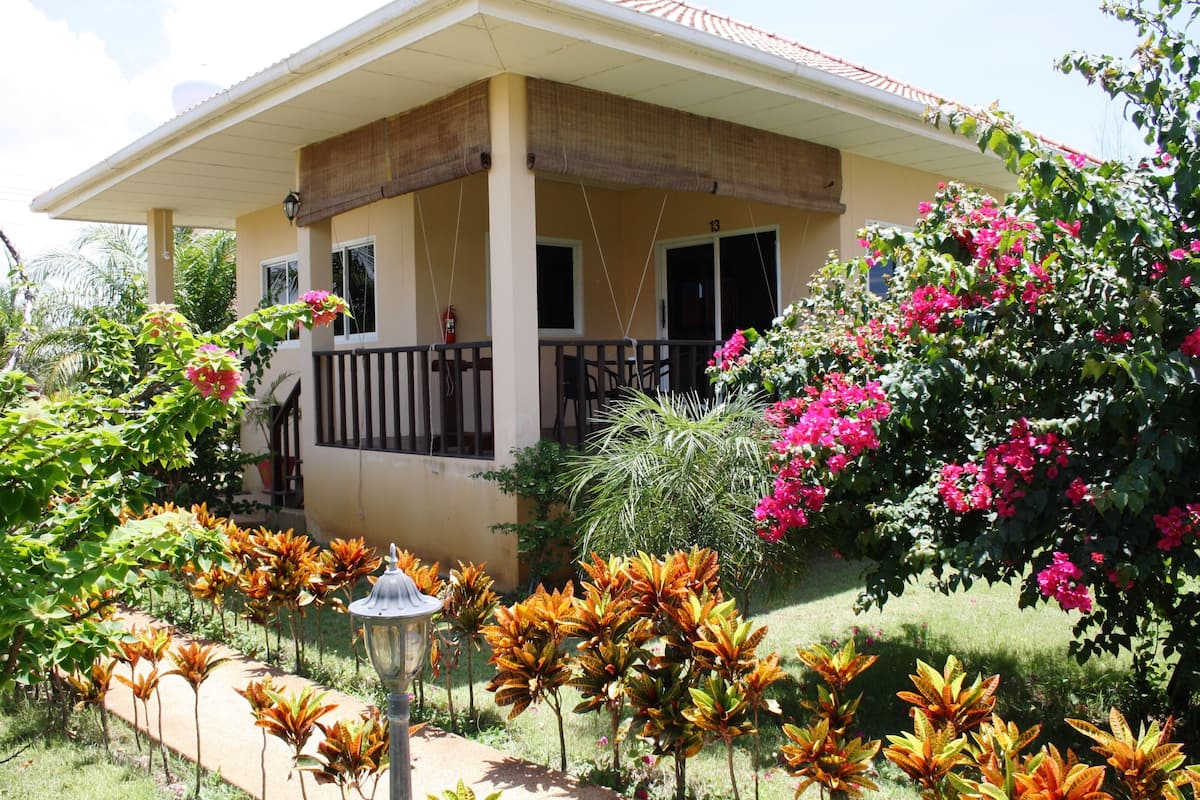Ein Haus mit überdachter Veranda, der Hausnummer '13' und einem Garten mit verschiedenen Pflanzen und Blumen.