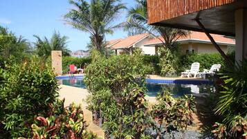 Zona de piscina con sillones de descanso y una piscina pequeña, rodeada de exuberante vegetación y un edificio con tejado de azulejos.