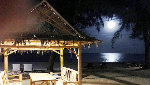 Una cabaña en la playa con techo de paja, muebles de madera y vista al océano por la noche.