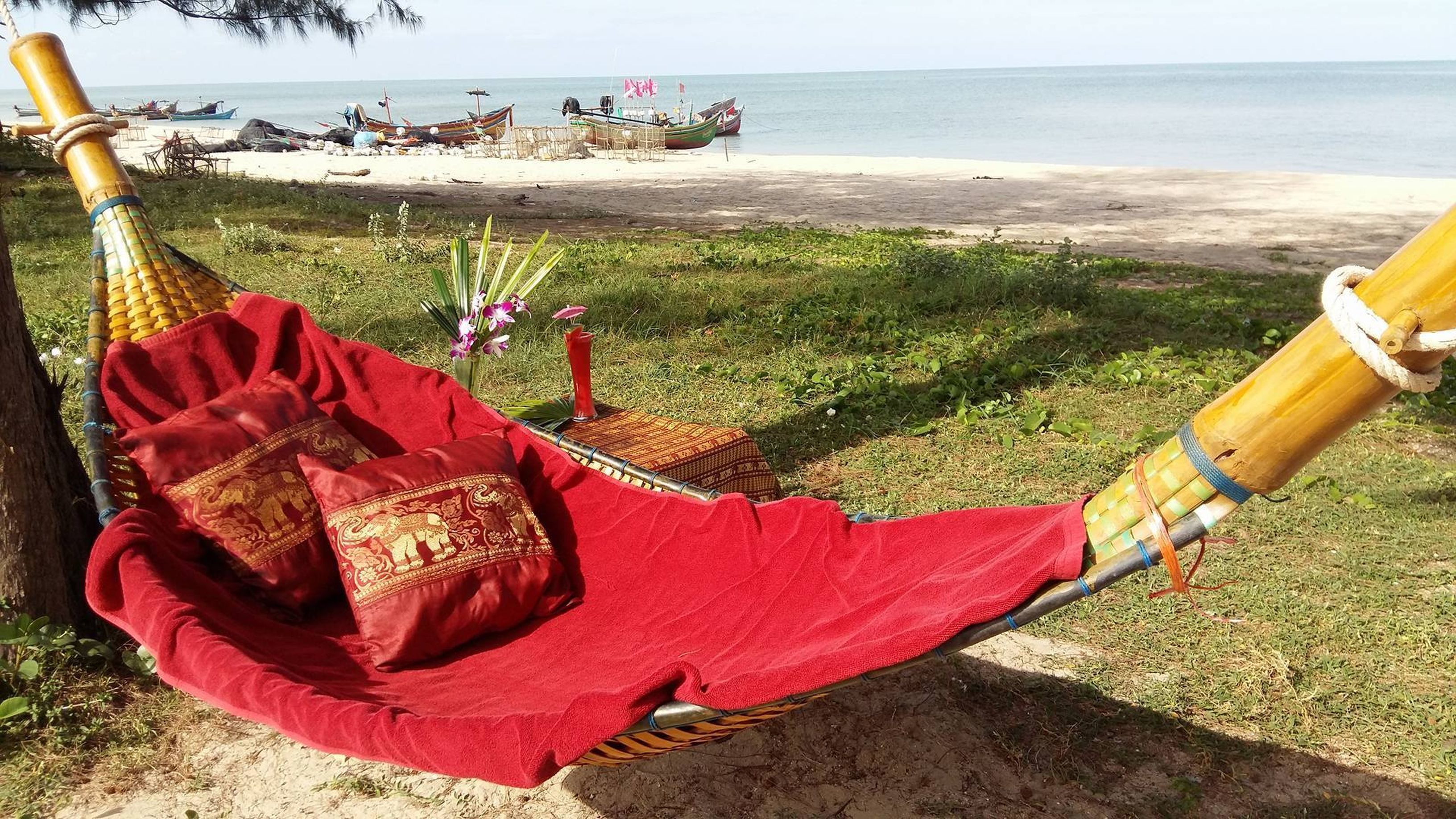 A hammock with a red blanket and pillows, set on a sandy beach with boats in the background.