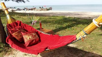 A hammock with a red blanket and pillows, set on a sandy beach with boats in the background.