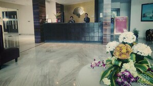A hotel reception area with a reception desk, two staff members, a decorative fan, and a floral arrangement. - The Legend Hotel (Prayagraj)
