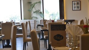 A restaurant with multiple tables and chairs, a large window with a view of greenery, and a sign with the restaurant's name. - The Legend Hotel (Prayagraj)