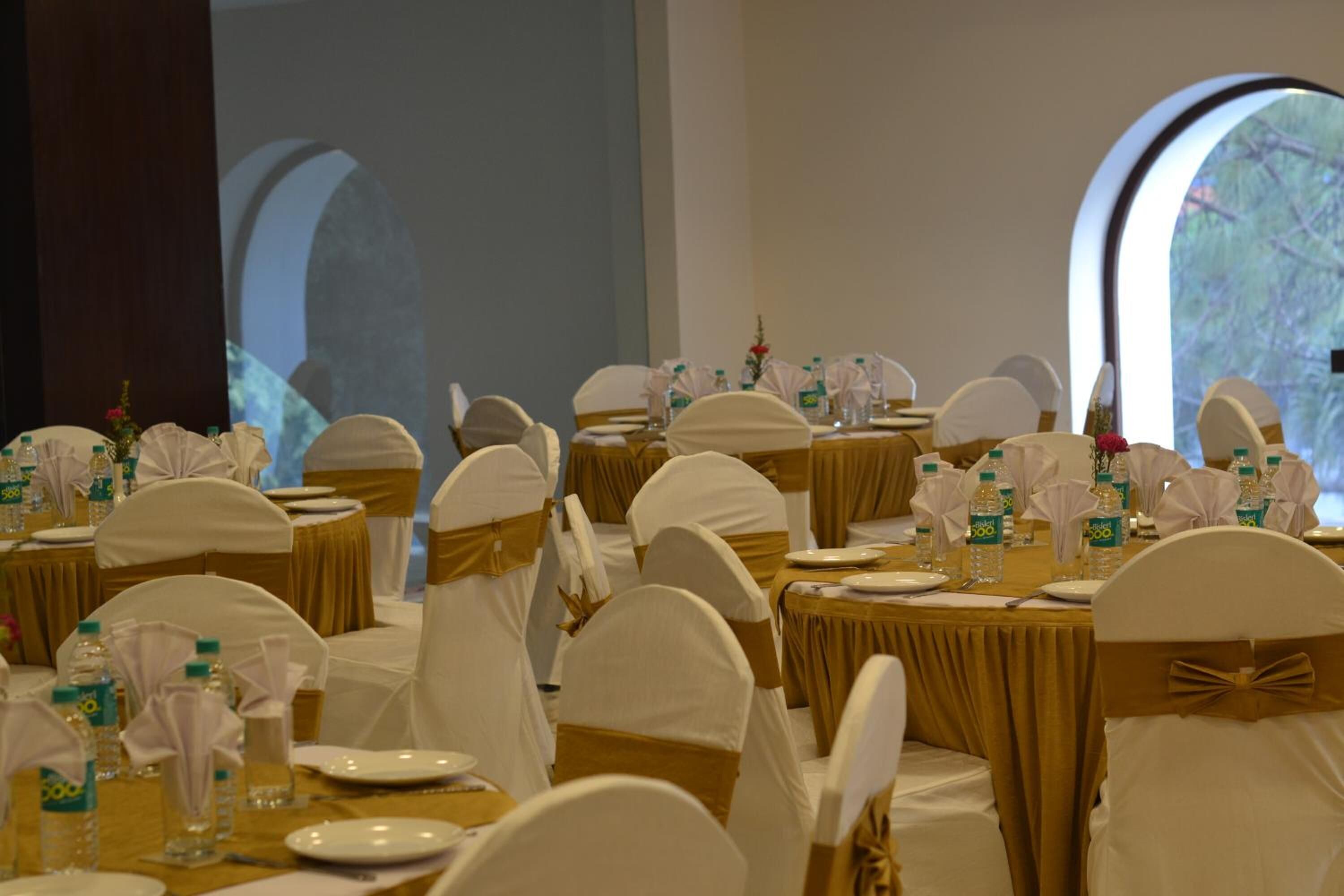 Une salle de banquet avec des tables rondes dressées pour un événement formel, agrémentée de chaises blanches, de nappes et de bouteilles d’eau.