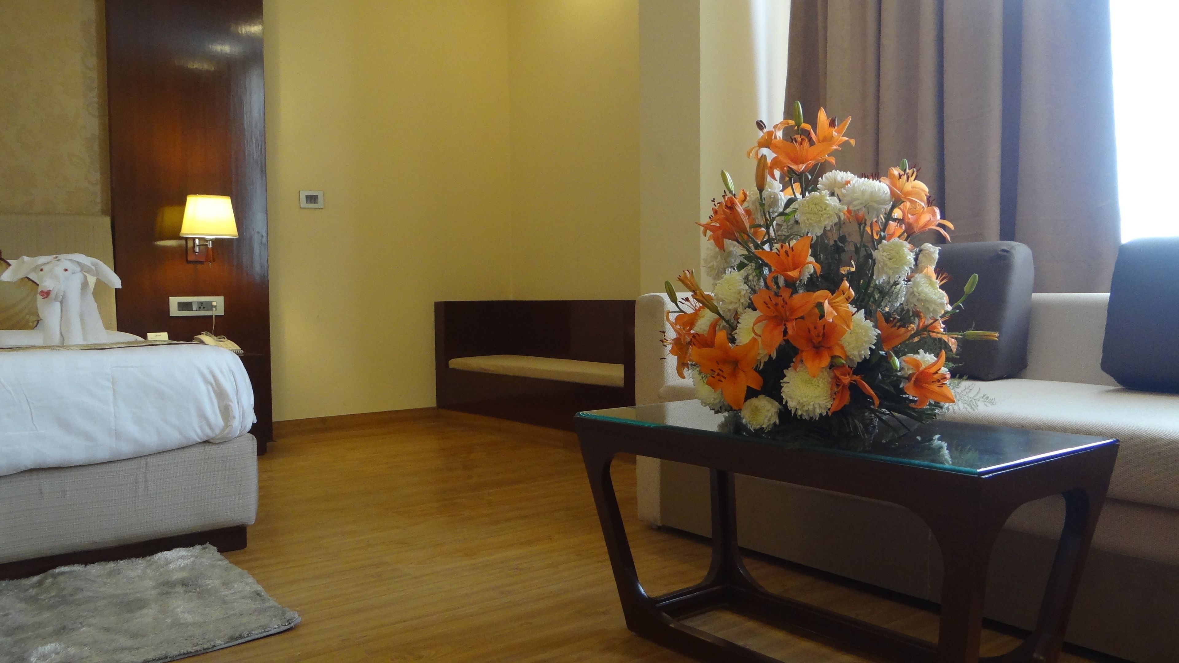 A hotel room with a bed, a sofa, a glass coffee table with a floral arrangement, and a lamp on the wall.