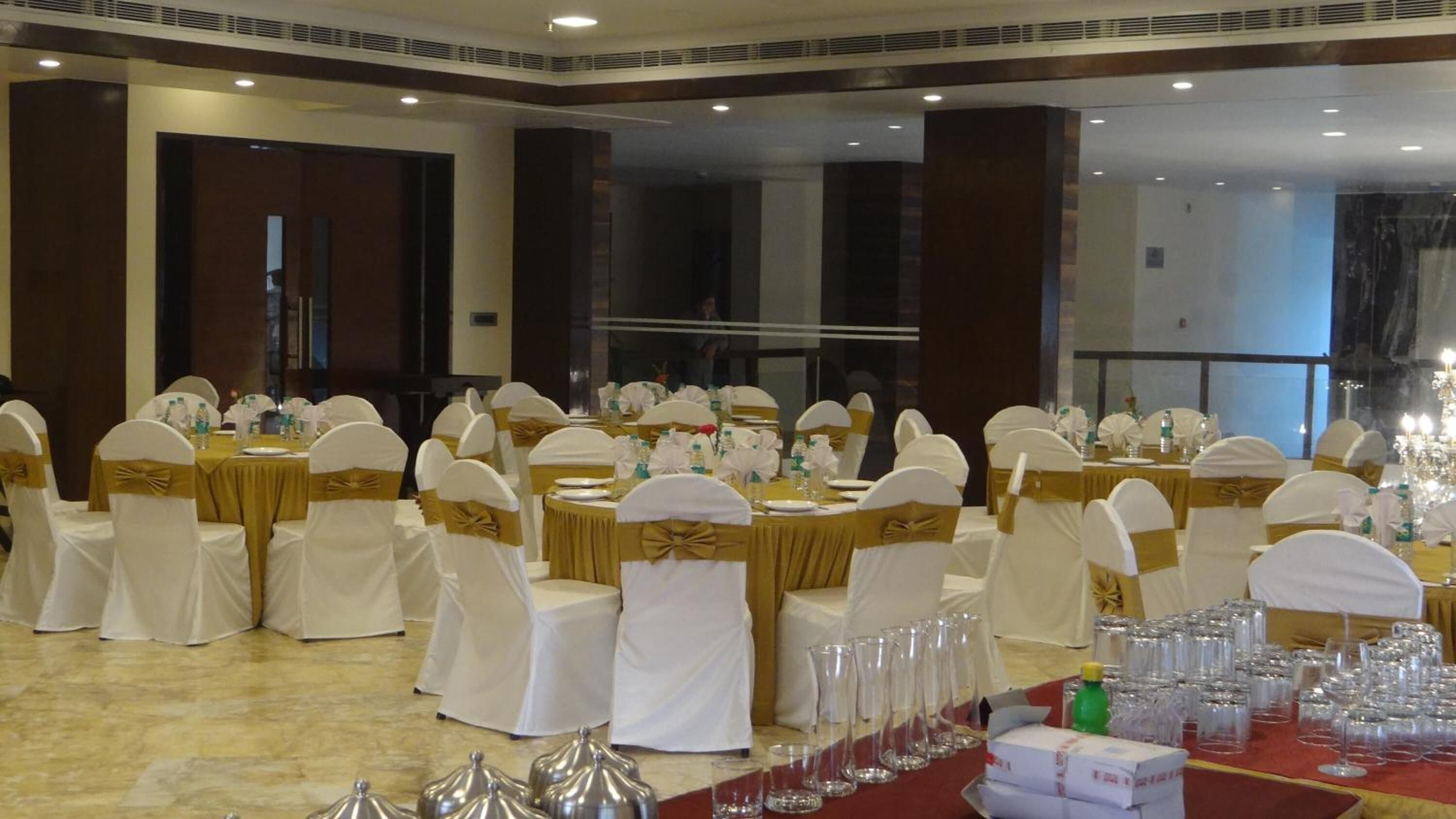 Un salón de banquetes con mesas redondas, sillas blancas y doradas, y cristalería transparente.