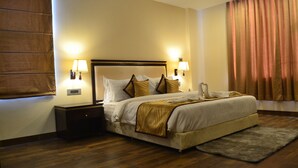 A neatly made bed with a headboard, two bedside tables with lamps, a wooden floor, and a window with curtains. - The Legend Hotel (Prayagraj)
