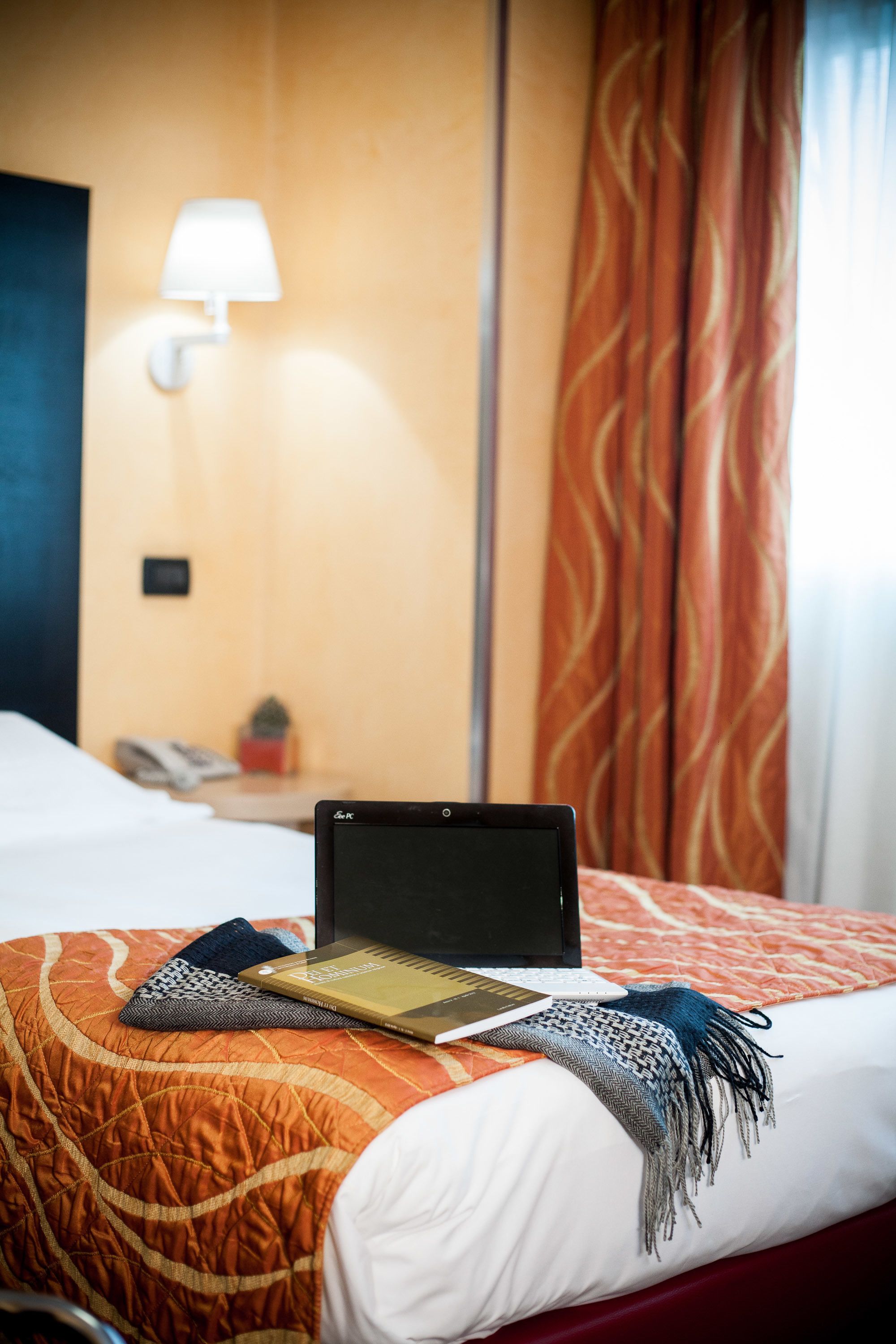 Una habitación de hotel con una cama, una computadora portátil, una lámpara y una mesita de noche con un teléfono y una pequeña planta.