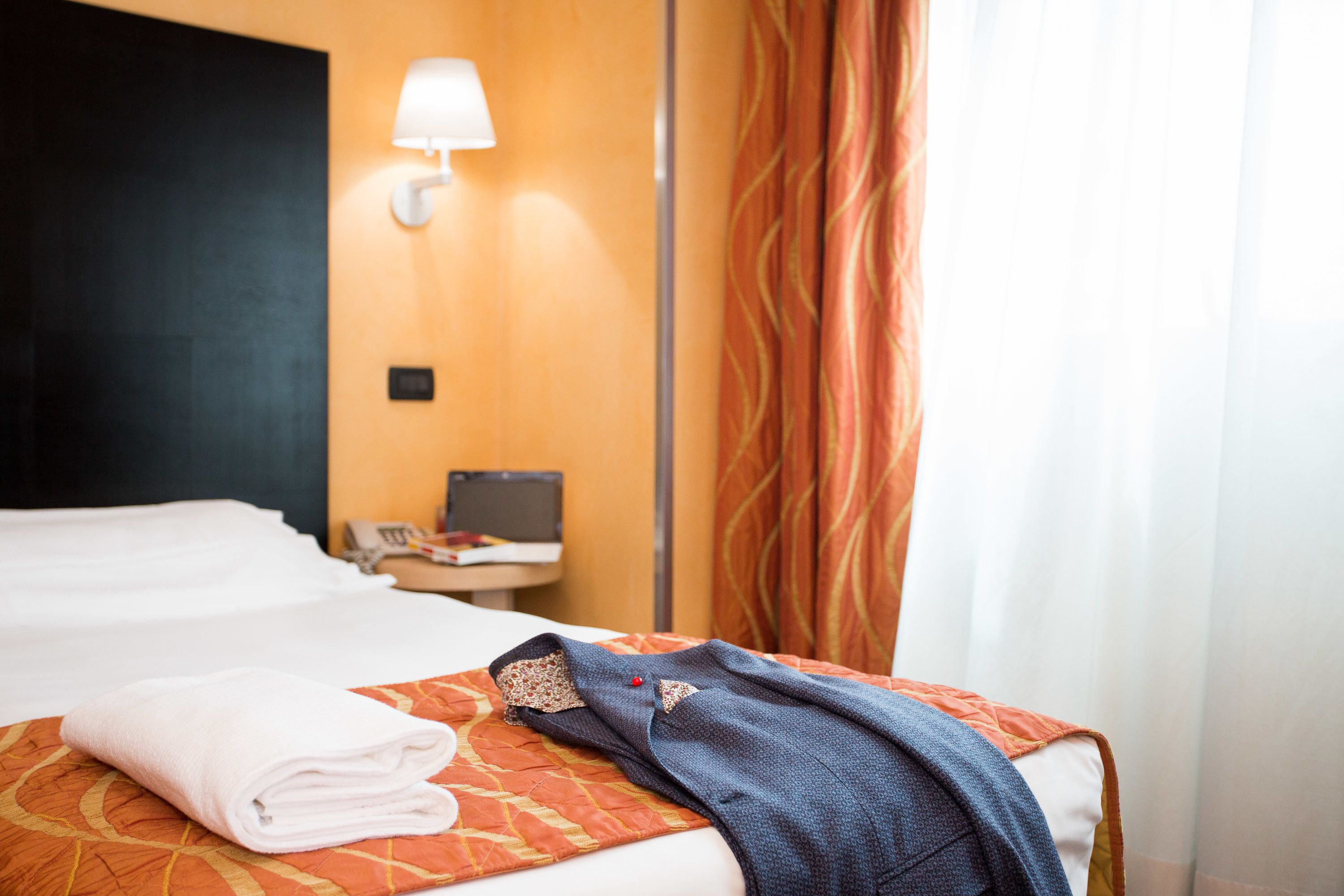 A neatly made bed with a folded towel, a blue blanket, and a patterned orange bedspread. There is a wall-mounted lamp and a small table with a phone and a book.