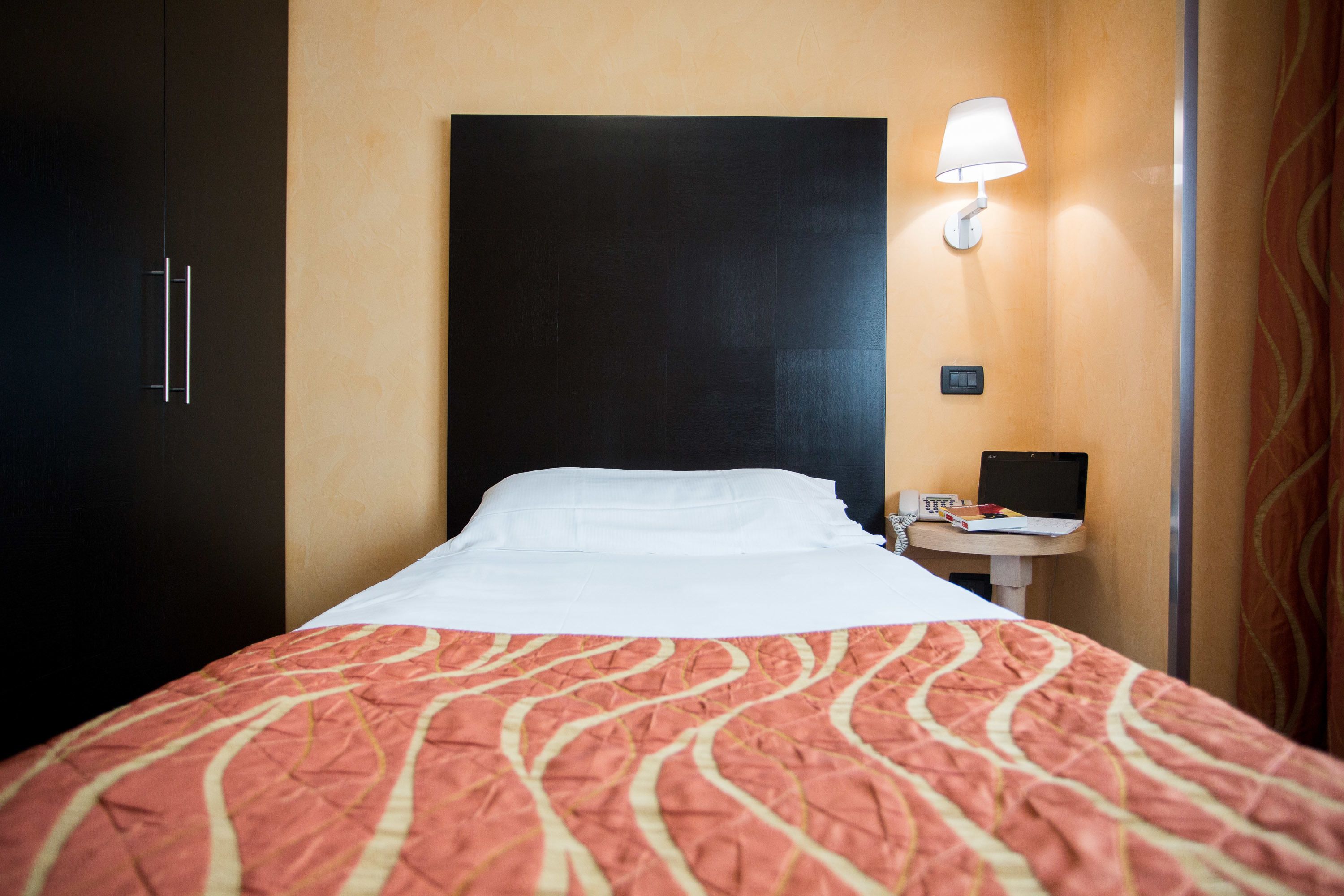 A hotel room with a bed, a bedside table with a lamp, a laptop, and a phone.
