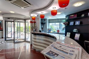 A modern hotel lobby with a curved reception desk, glass doors leading outside, and a digital clock displaying the time. - Hotel Majorana (Rende)