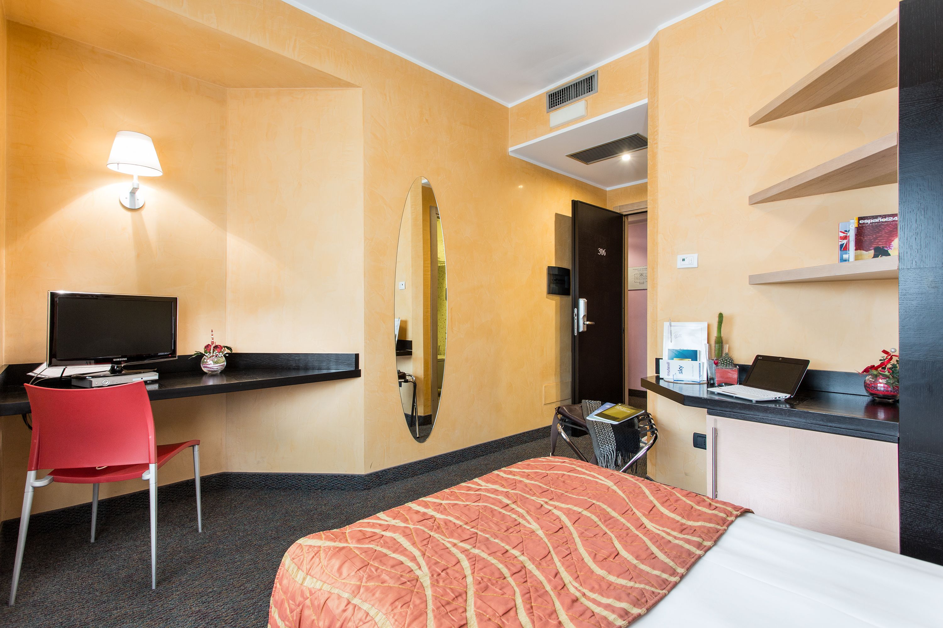 Una habitación de hotel con una cama, un escritorio con una computadora, una silla, un espejo y una estantería con objetos.
