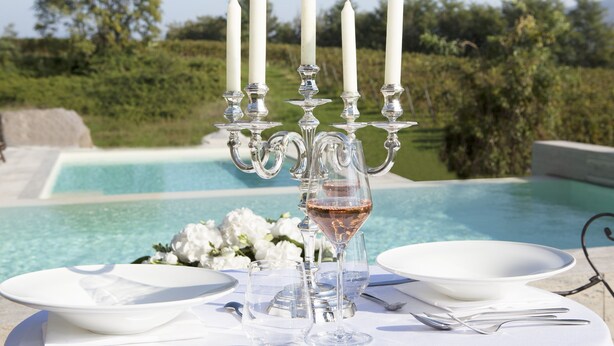 A table set for two with white plates, silverware, and a glass of rosé wine. A candelabra with white candles is in the center. The table is outdoors with a pool and greenery in the background. - Podere Castel Merlo Resort (Villongo)