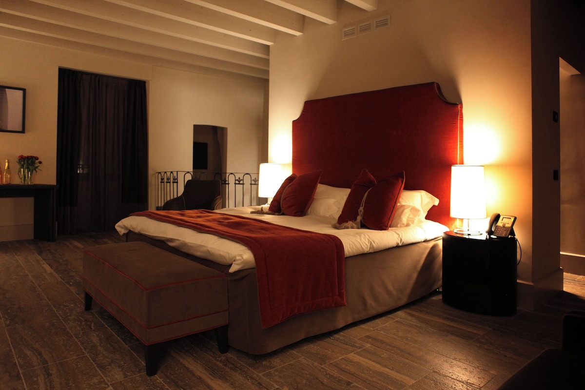 Un dormitorio con una cama grande, una mesita de noche con una lámpara y una pequeña mesa con un jarrón y flores.