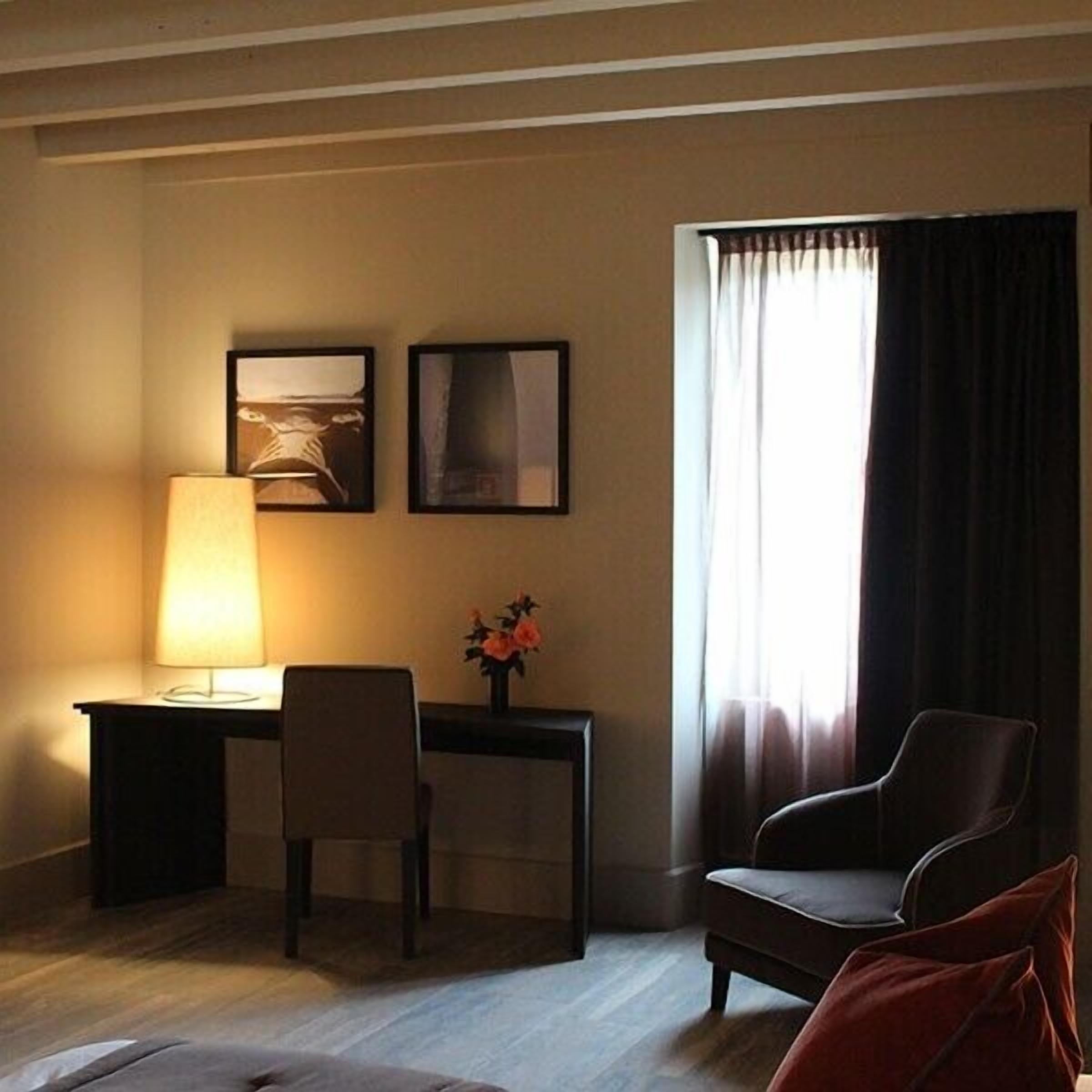 A hotel room with a desk, two chairs, a lamp, a vase with flowers, and a large window with curtains.