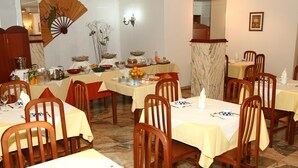 A dining area with multiple tables, chairs, and a fan. Tables are covered with yellow tablecloths, and there are various dishes and utensils on them. - Hotel JR (Sanxenxo)
