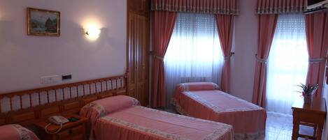 A hotel room with two single beds, a wooden headboard, a bedside table with a telephone, and a small table with a plant.