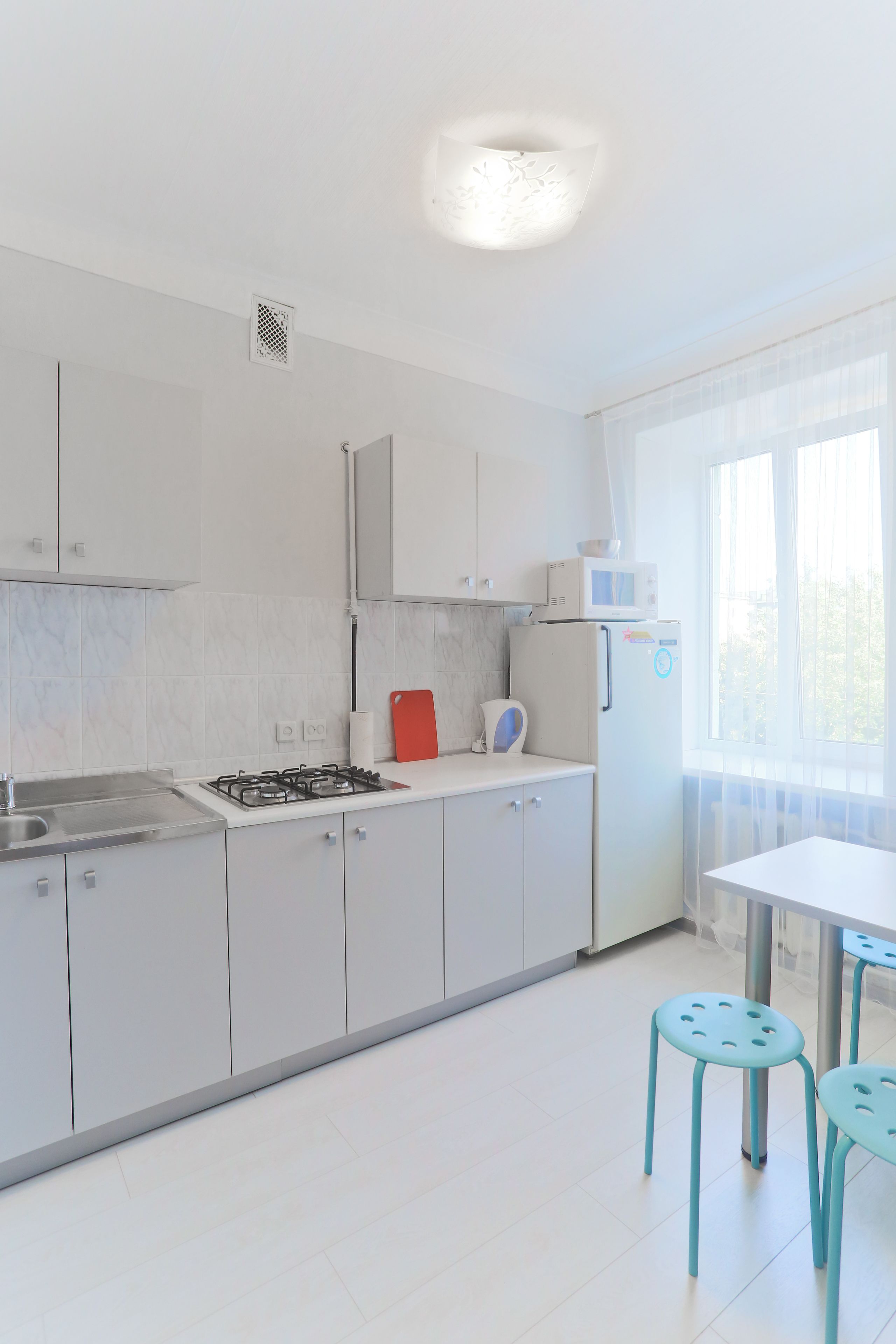 Una cocina moderna con armarios blancos, estufa a gas, microondas y refrigerador. Hay dos taburetes azules y una tabla de cortar roja.