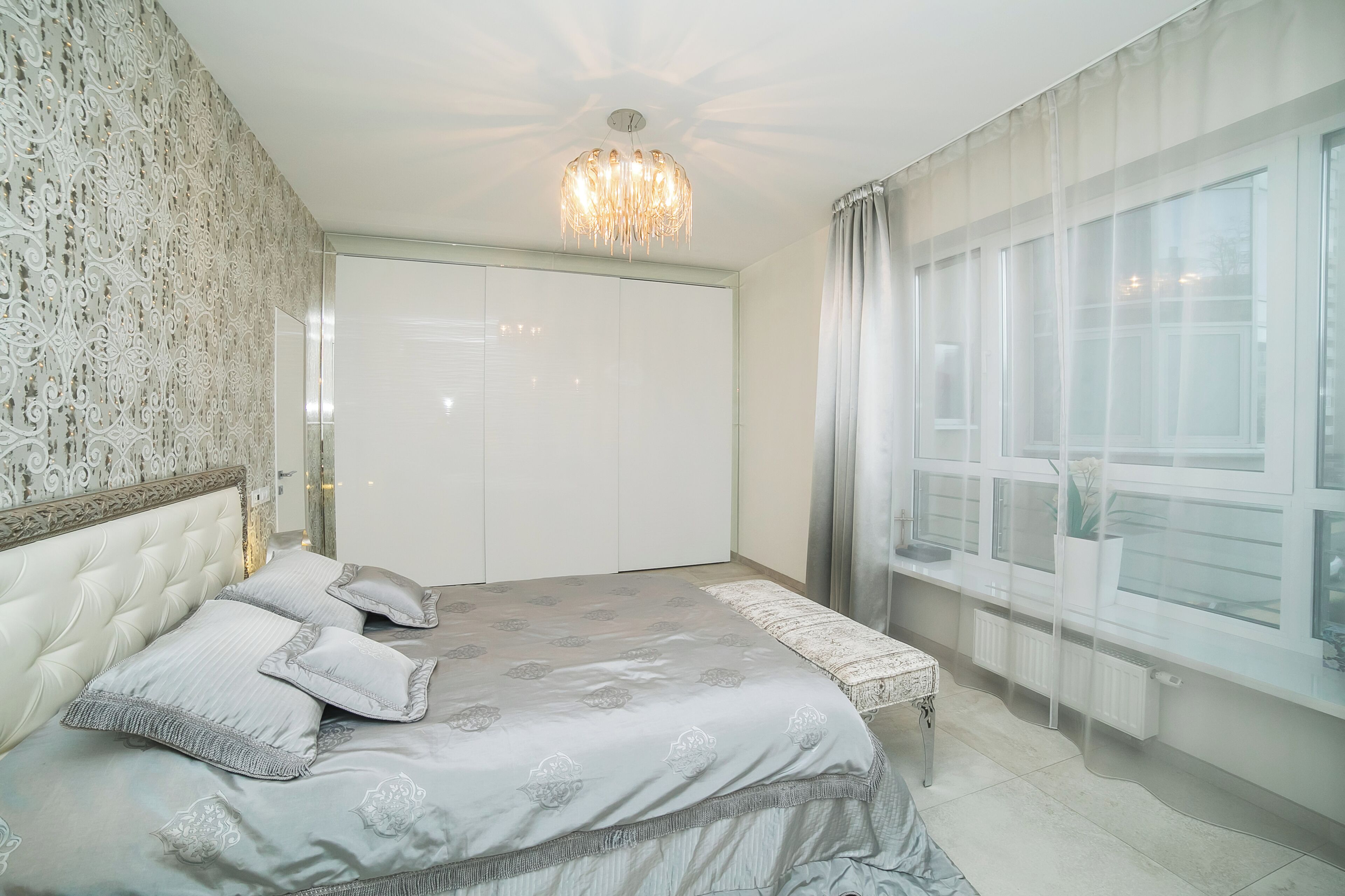 A bedroom with a large bed, a chandelier, a wardrobe, and a window with curtains.