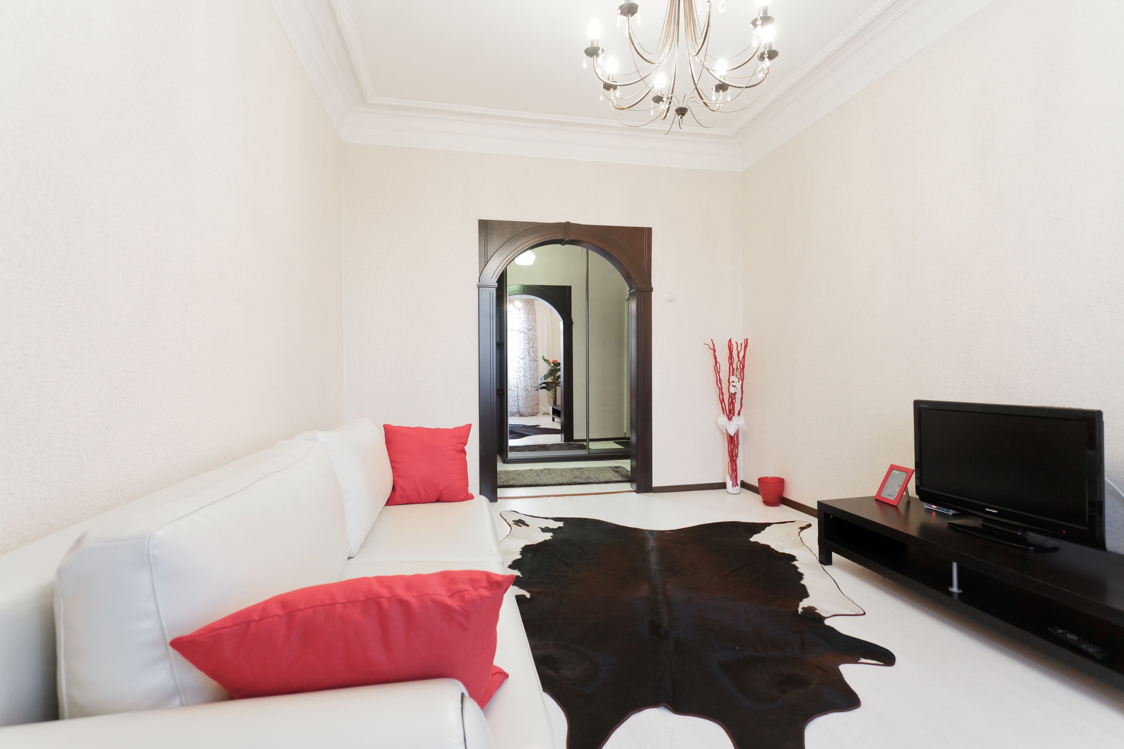 A modern living room with a white sofa, red cushions, a black leather rug, a television on a black stand, and a chandelier.