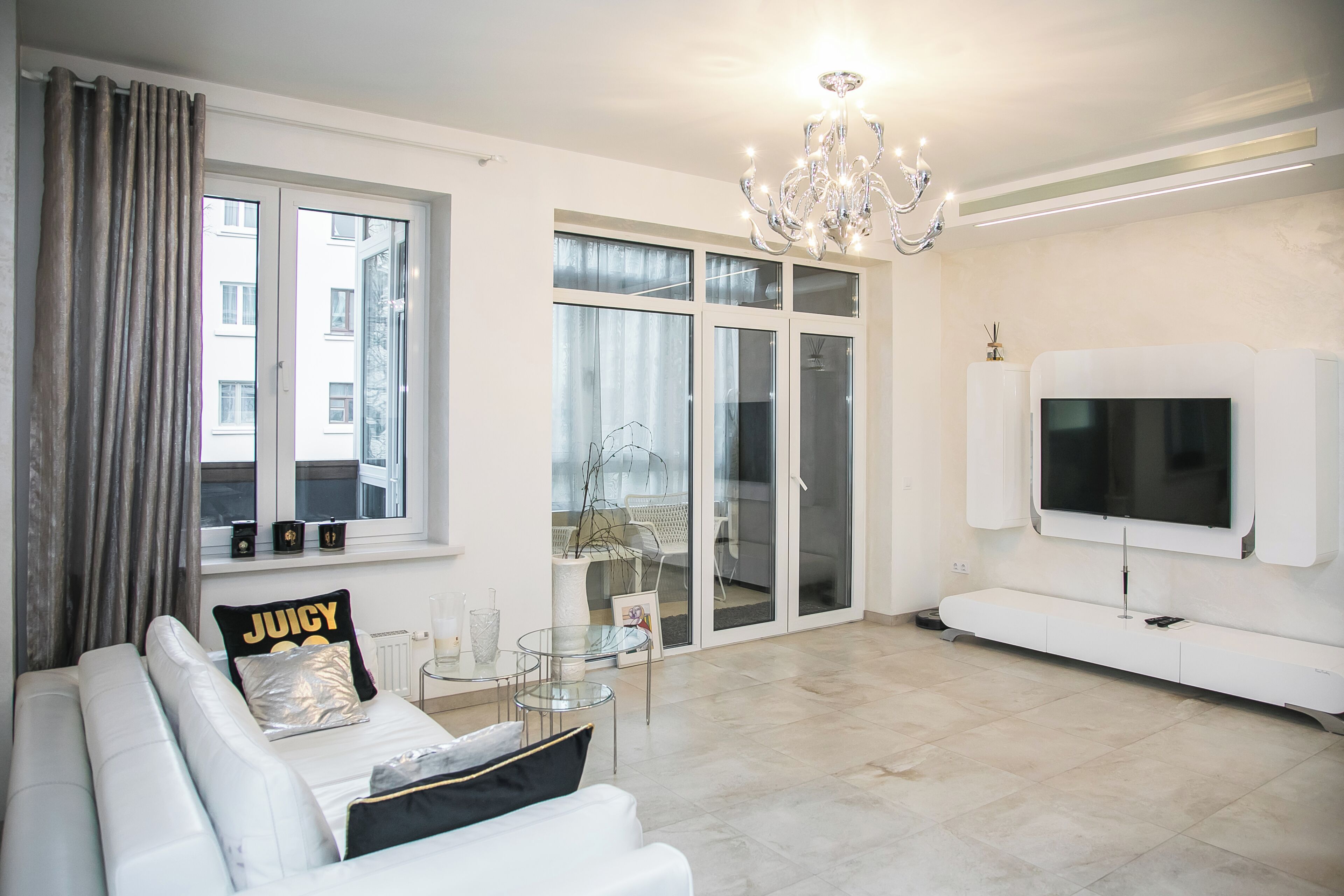 Een moderne woonkamer met een witte bank, een glazen salontafel, een kroonluchter, een flatscreen televisie en een schuifpui naar een balkon.