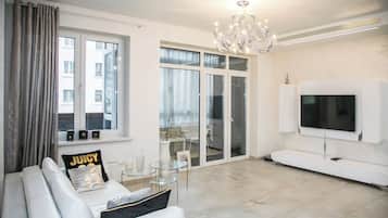 Una sala de estar moderna con un sofá blanco, una mesa de centro de vidrio, una araña, un televisor de pantalla plana y un puerta corredera de vidrio que da a un balcón.
