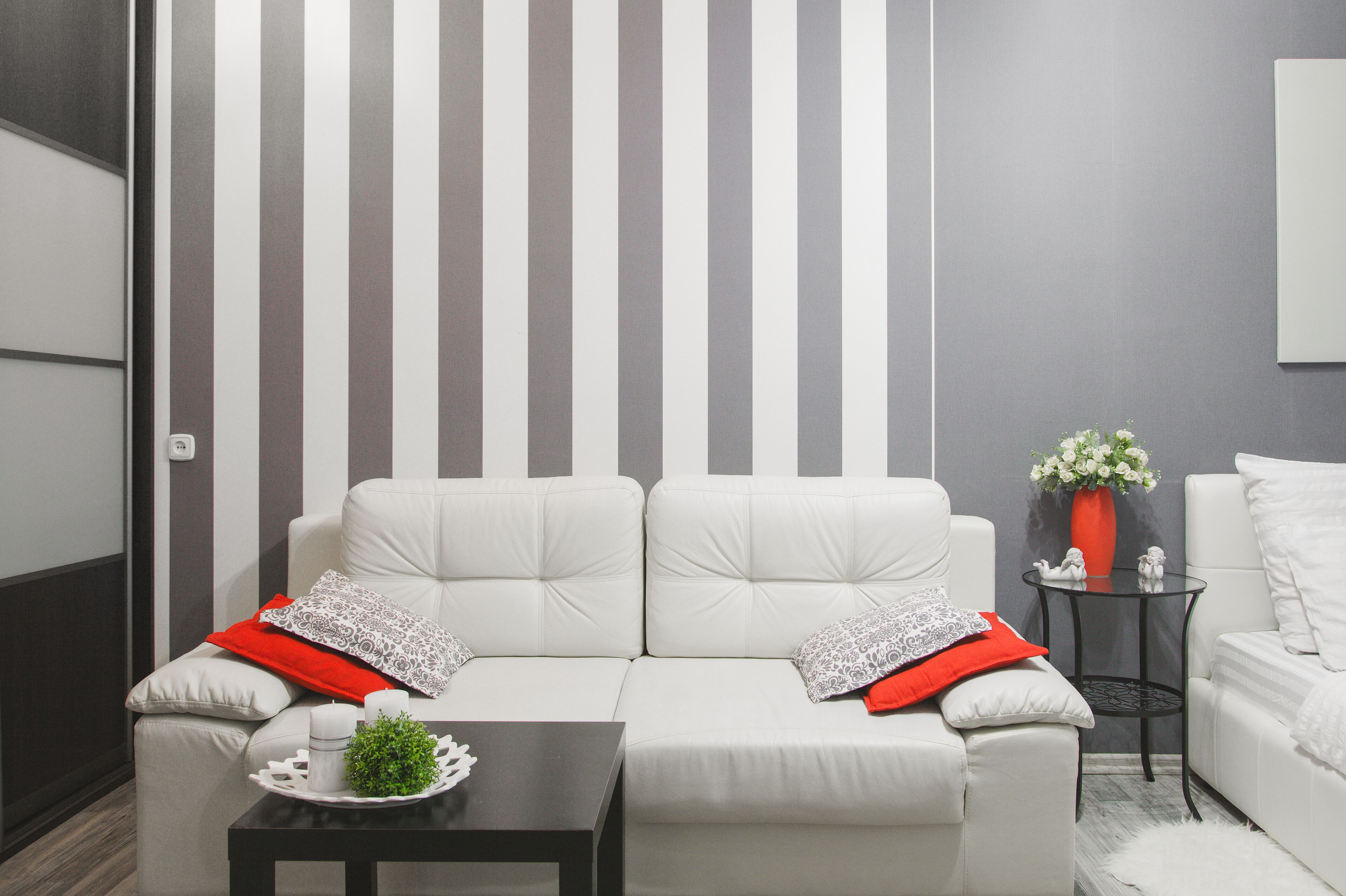 A modern living room with a striped wall, a white sofa set, a small table with a tray, and a vase with flowers.