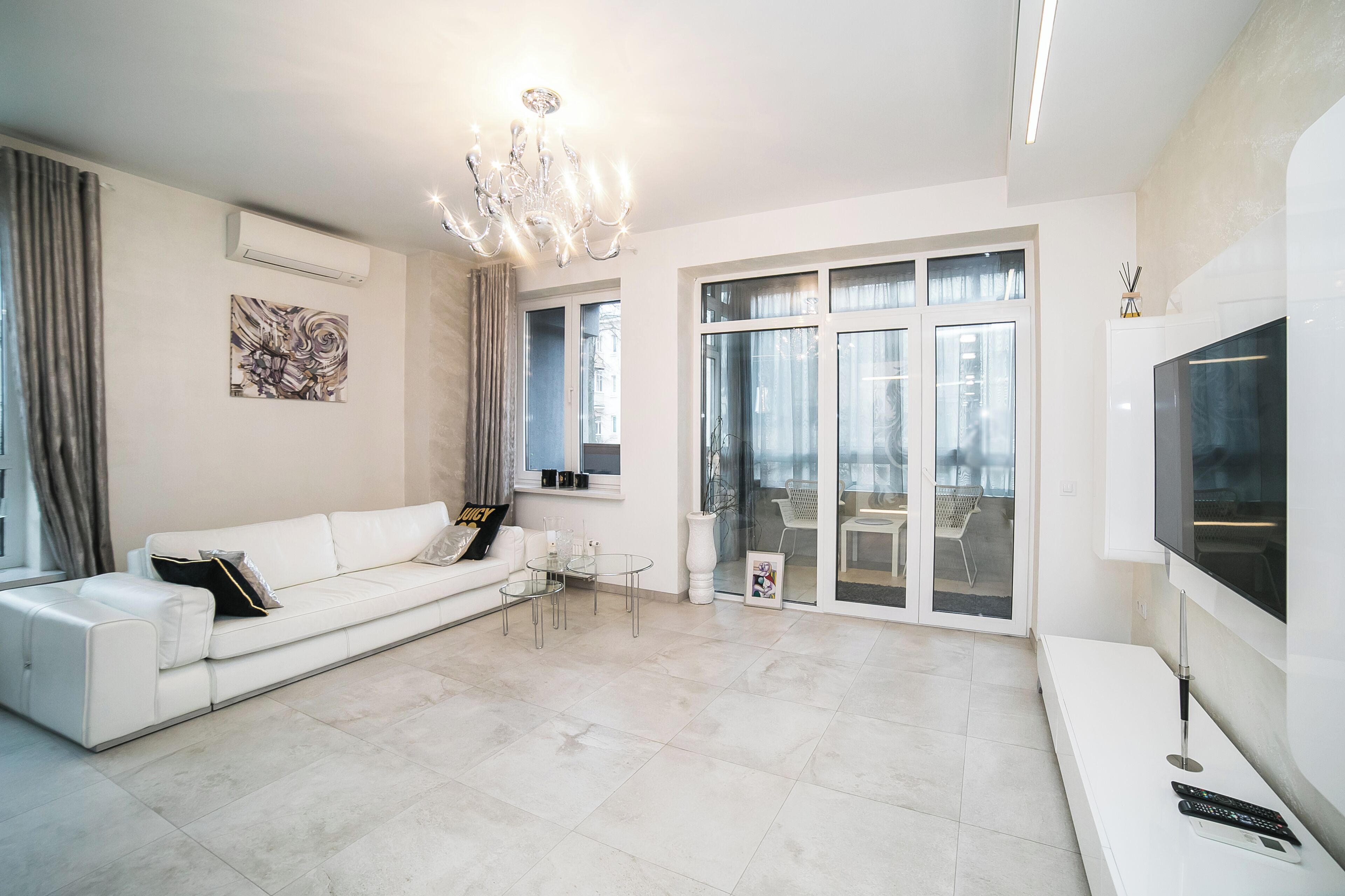 A modern living room with a white sofa, a glass coffee table, a chandelier, and a large TV.