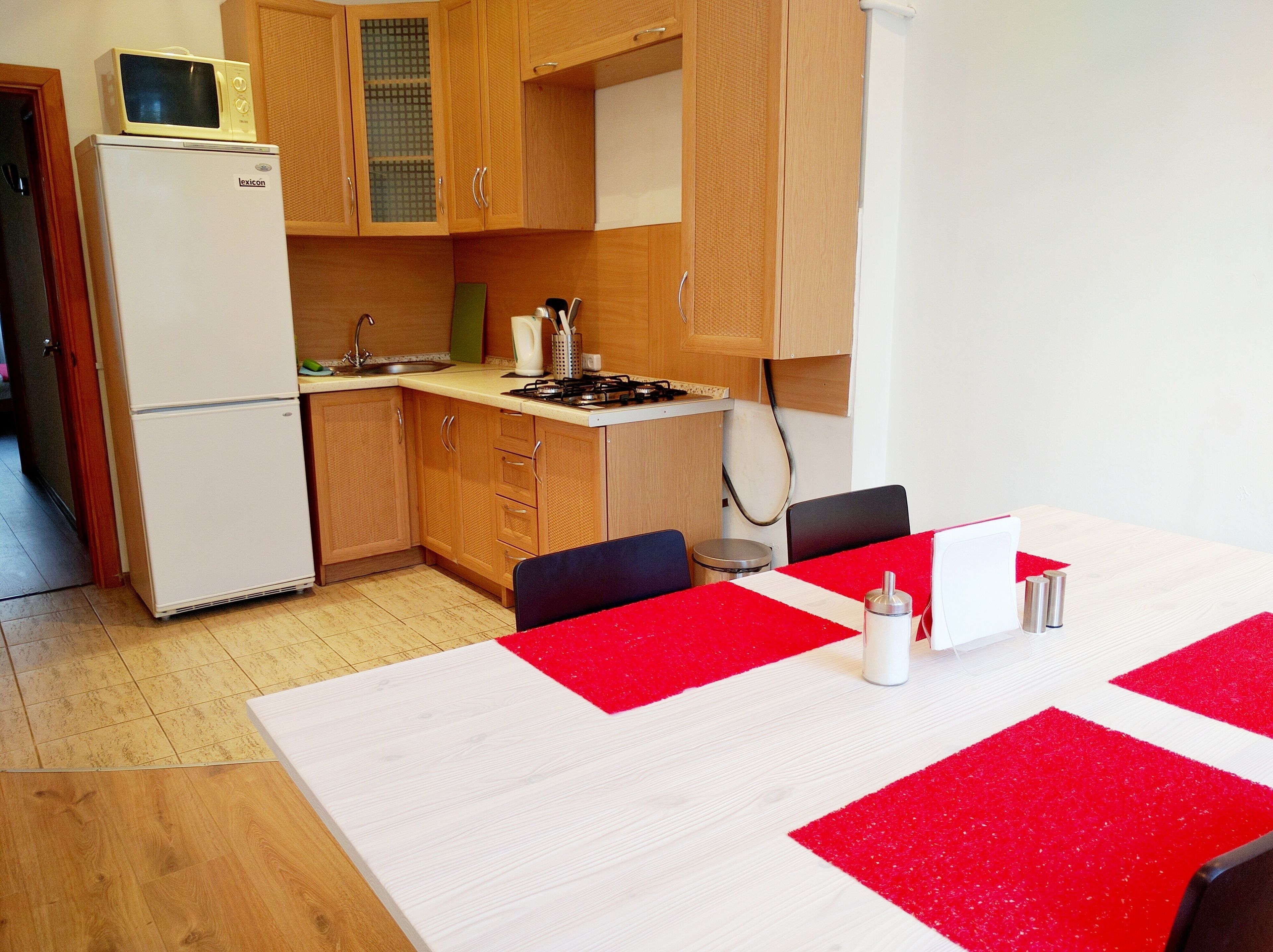 Una cucina compatta con mobili in legno, un frigorifero bianco, un forno a microonde, un piano cottura e un tavolo da pranzo con sottopiatti rossi.