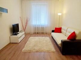 Ein Wohnzimmer mit einer Couch, einem Fernseher auf einem Schrank, einer Stehlampe und einem Fenster mit Vorhängen.