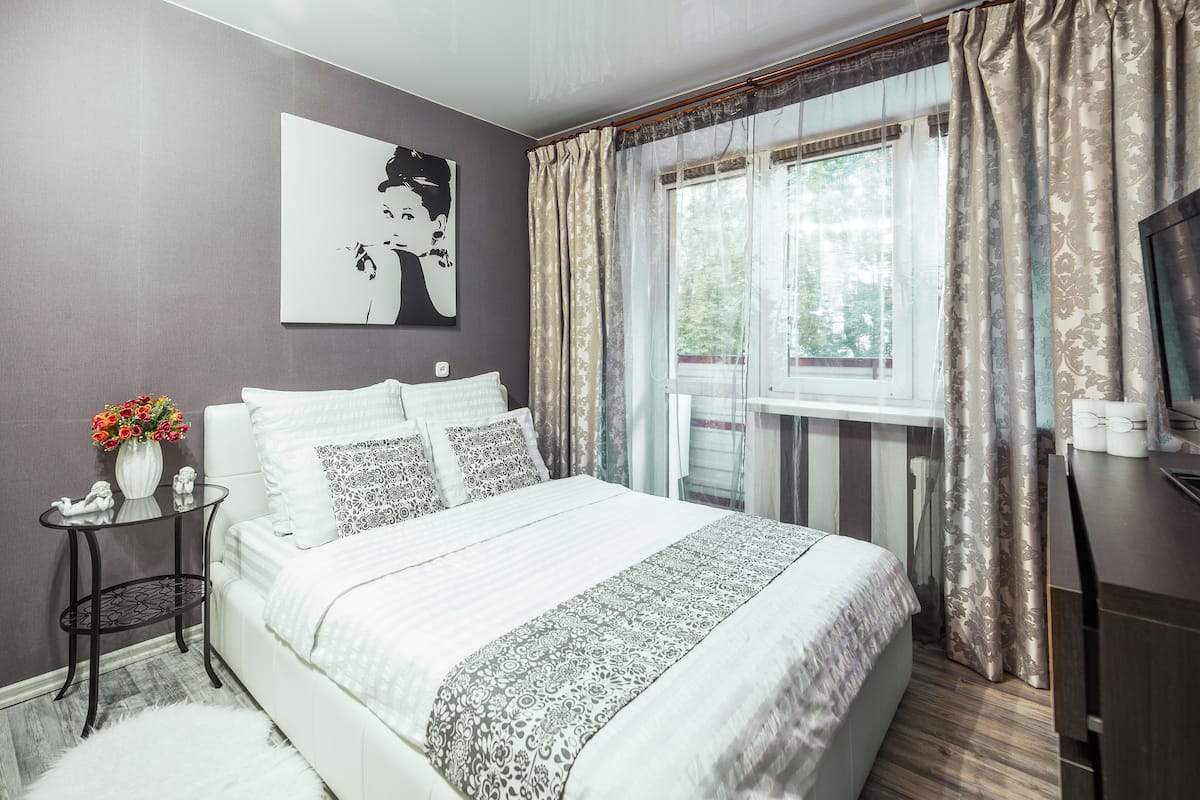 Une chambre à coucher bien rangée, avec un lit, une table de chevet ornée d’un vase de fleurs, un tableau accroché au mur et une fenêtre avec des rideaux.